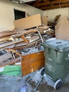 Cluttered garage filled with debris and a green trash bin in the corner.