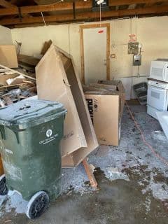 Cluttered garage interior with cardboard boxes, trash can, and appliances in disarray.