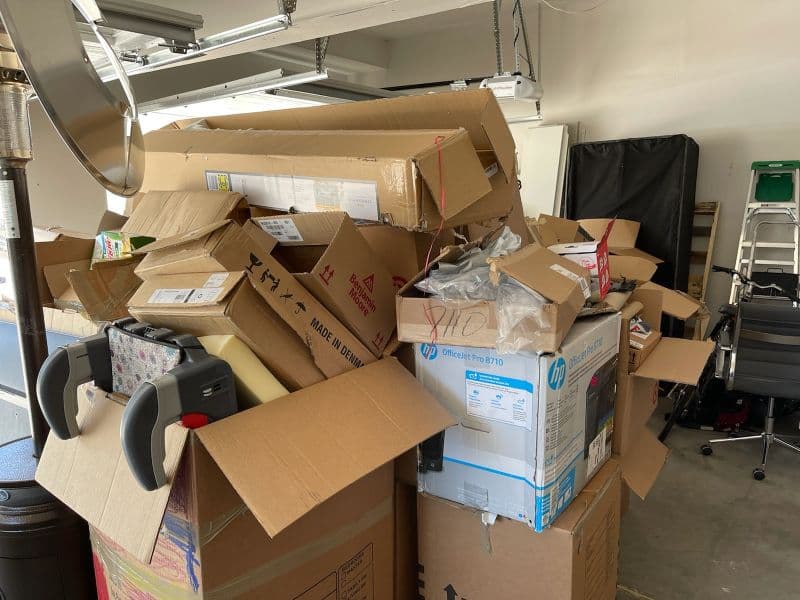 Stack of large cardboard boxes piled high in a garage, surrounded by various items.