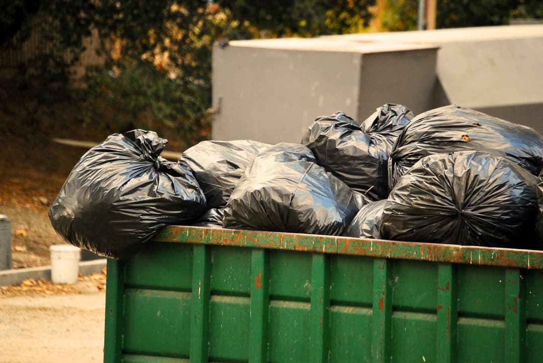 Green dumpster filled with black trash bags in an outdoor setting.