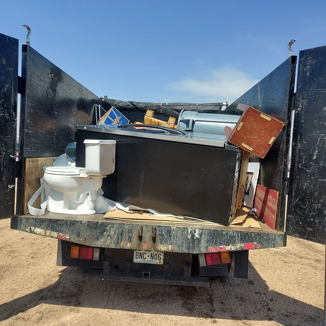 Truck bed filled with various discarded appliances and furniture, including a toilet and refrigerator.